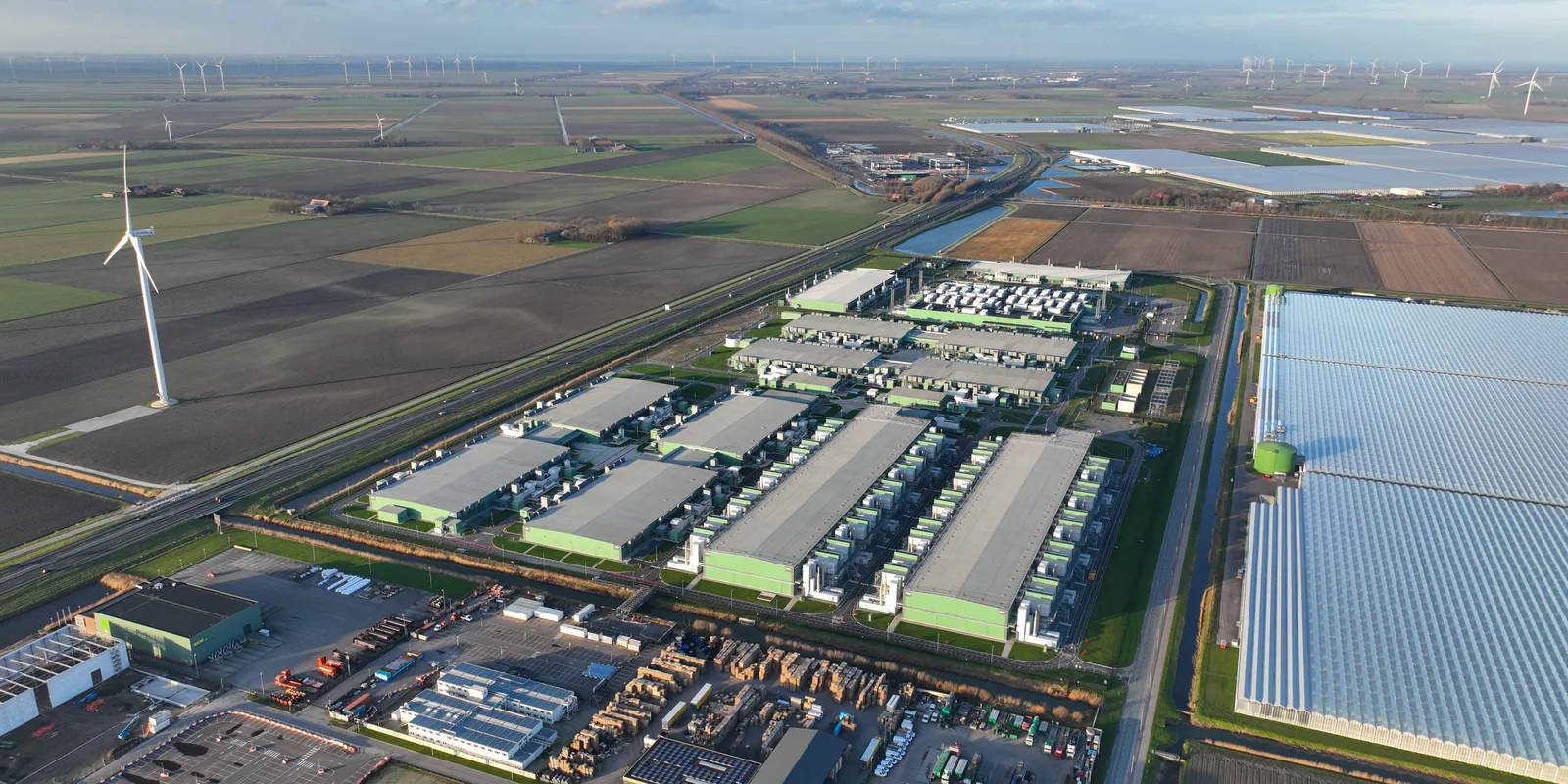 Aerial view of a large data center complex wedged into rural farmland, flanked by wind turbines, drainage canals, and greenhouses.